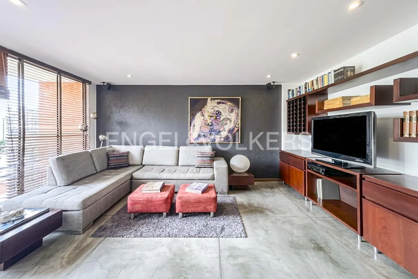 Living room with gray sectional sofa, red ottomans, and a dark gray accent wall with colorful art. A TV and shelves are on the right.