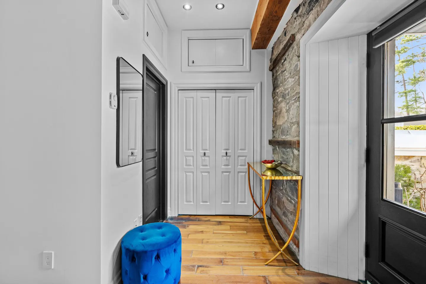 Entryway with white walls, wood floors, and a blue ottoman. A gold console table sits near a stone wall and black door.