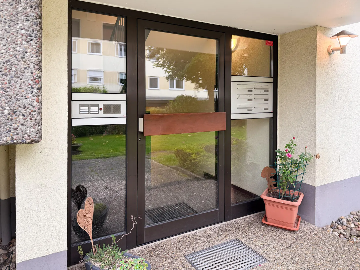 Apartment building entrance with a dark brown framed glass door, mailboxes, doorbell, and potted plants on the concrete porch.