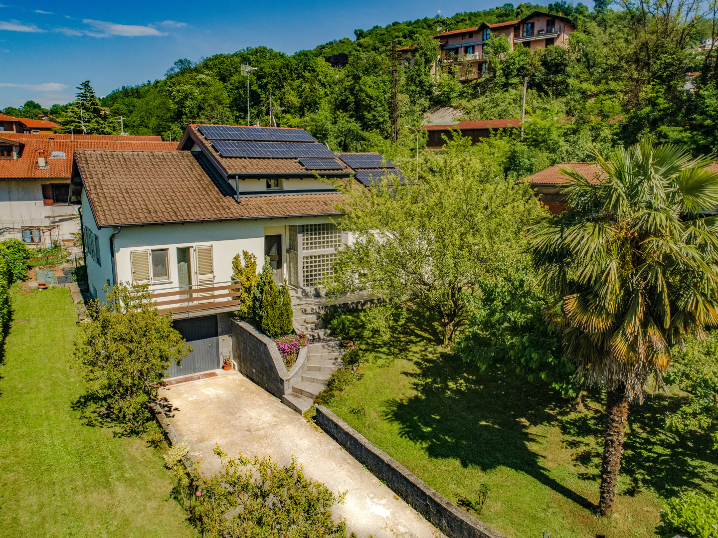 A two-story house with solar panels on a brown roof, a balcony, and a driveway, set in a lush, green hillside landscape.