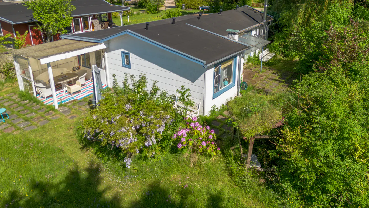 Aerial view of a white cottage with a black roof, a covered patio with outdoor furniture, and a lush green garden.