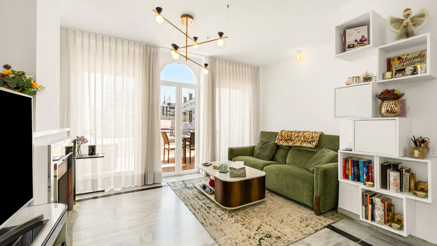 Bright living room with green sofa, marble floors, and white walls. A modern chandelier hangs above a coffee table. Balcony doors let in natural light.