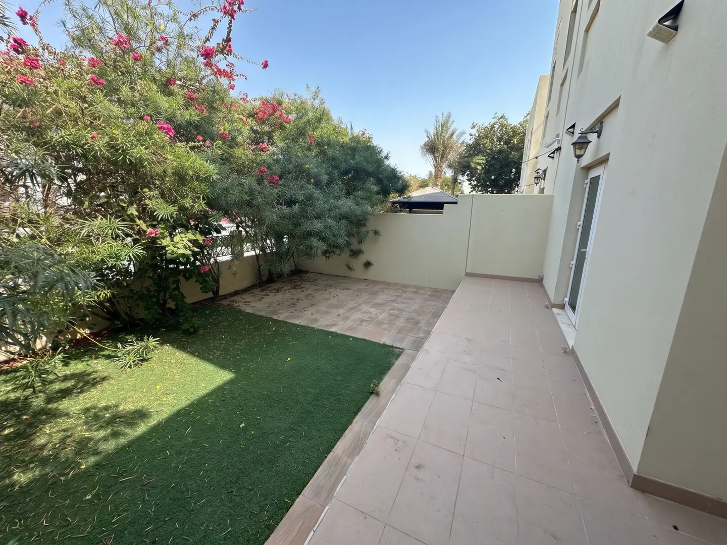 A backyard with green grass, trees with pink flowers, and a tiled patio next to a light beige building.