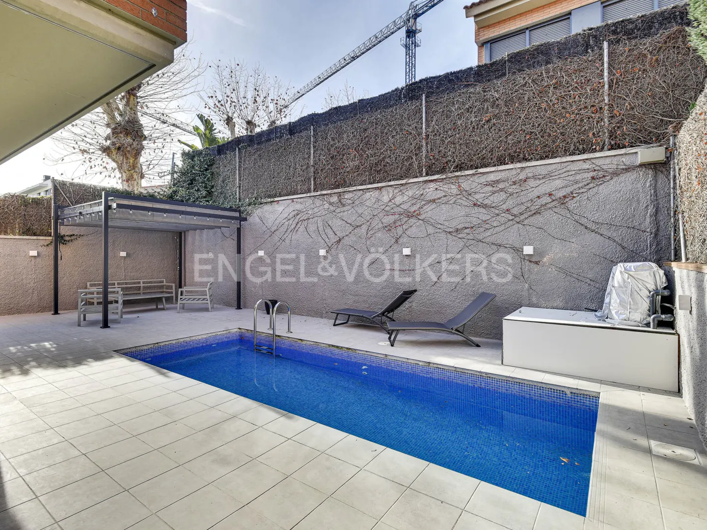 A backyard with a blue tiled pool, two lounge chairs, a pergola with benches, and a grey wall with vines.