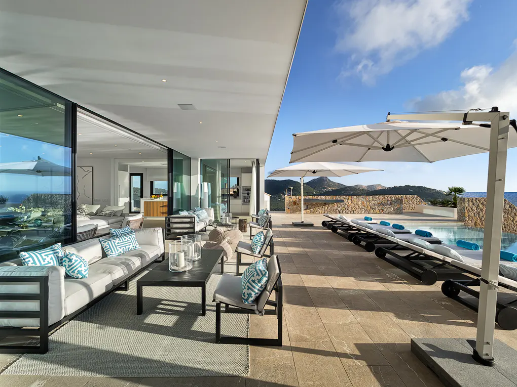 Luxury villa patio with white sofas, chairs, and sun loungers. A pool and ocean view are visible under a blue sky.
