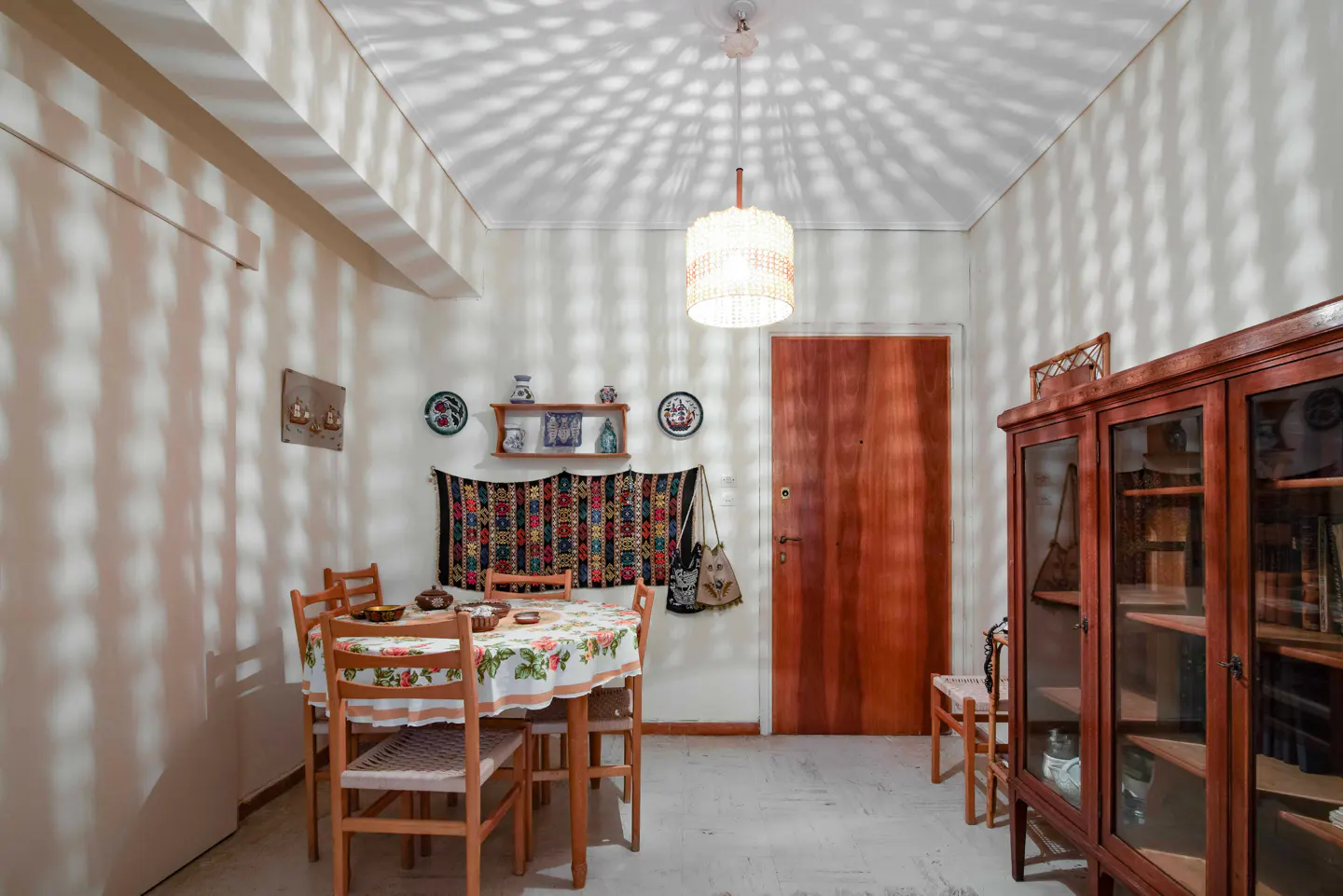A dining room with a table, chairs, a wooden cabinet, and a door. The walls are white, and the ceiling has a patterned light effect.