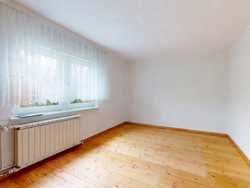 Empty room with wood floors, white walls, and a radiator under a window with sheer white curtains.