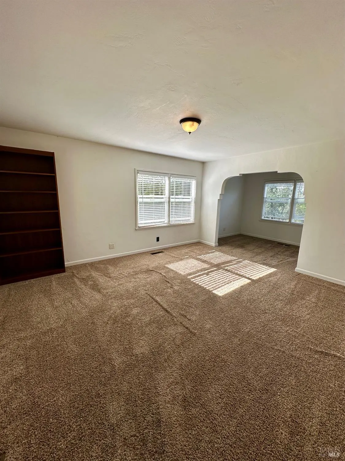 A bright, empty living room with brown carpet, white walls, and sunlight streaming through the windows. A dark bookcase stands against one wall.