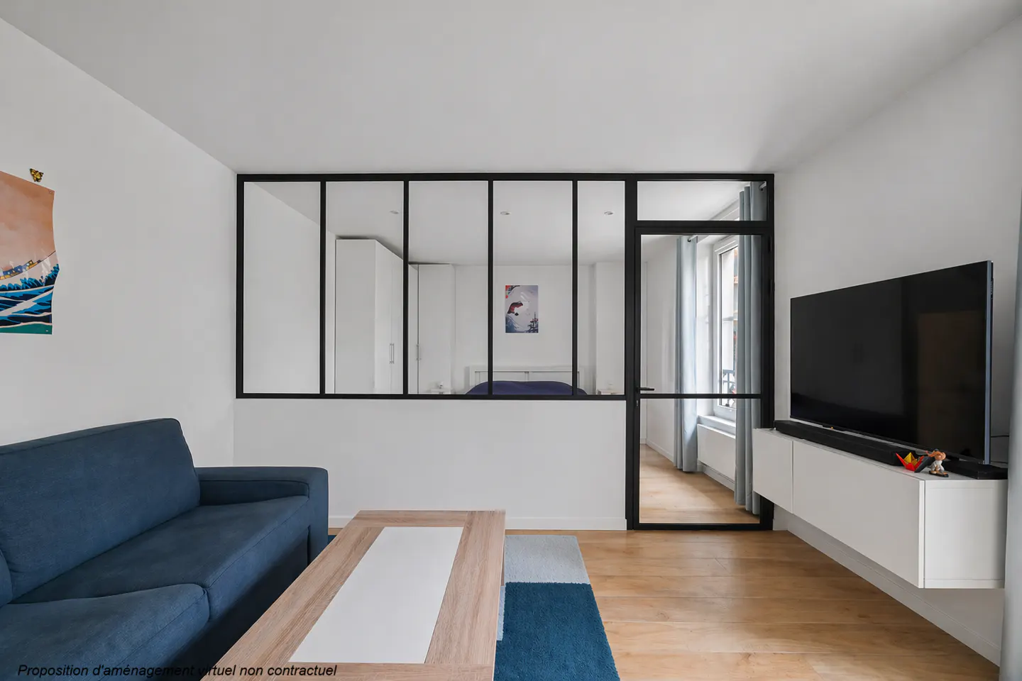 Living room with blue sofa, wood floors, and a black framed glass wall separating the bedroom. A TV is mounted on a white cabinet.