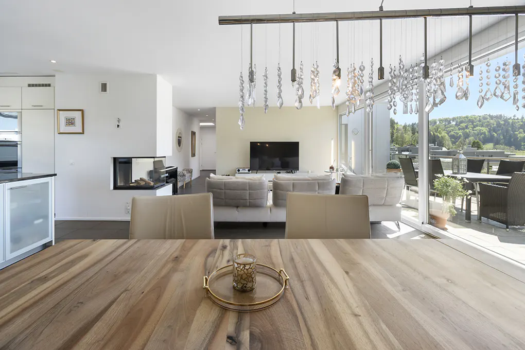 Open-concept living space with a wood dining table, crystal chandelier, and a view of a balcony and green hills.