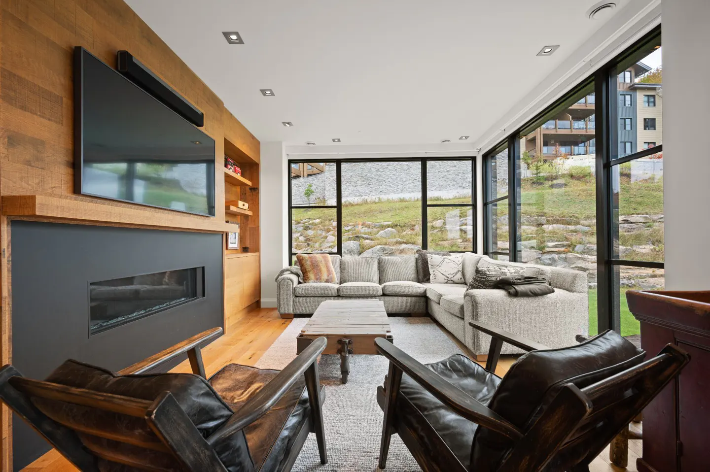 Living room with a gray sectional, leather chairs, fireplace, and large windows overlooking a grassy hill.