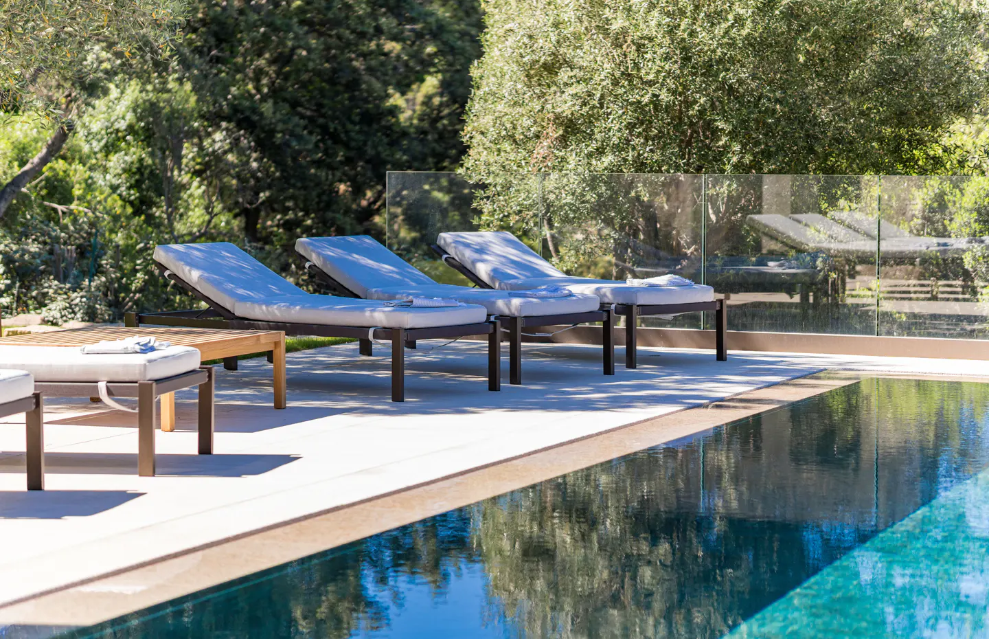 Three lounge chairs with white cushions sit on a sunny patio beside a swimming pool, surrounded by lush green trees.