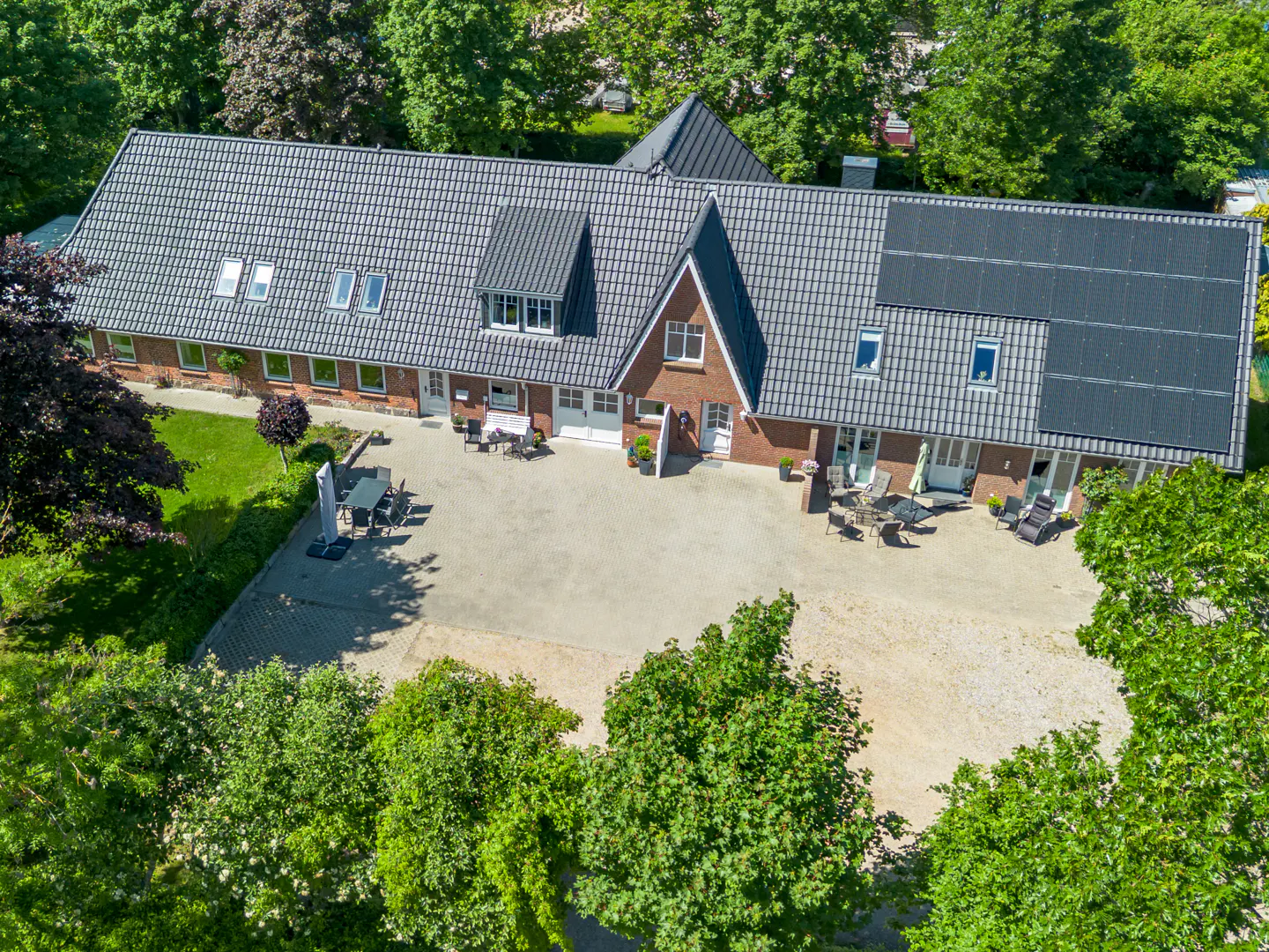 Aerial view of a long brick house with a dark tiled roof, solar panels, and a large paved courtyard with patio furniture.