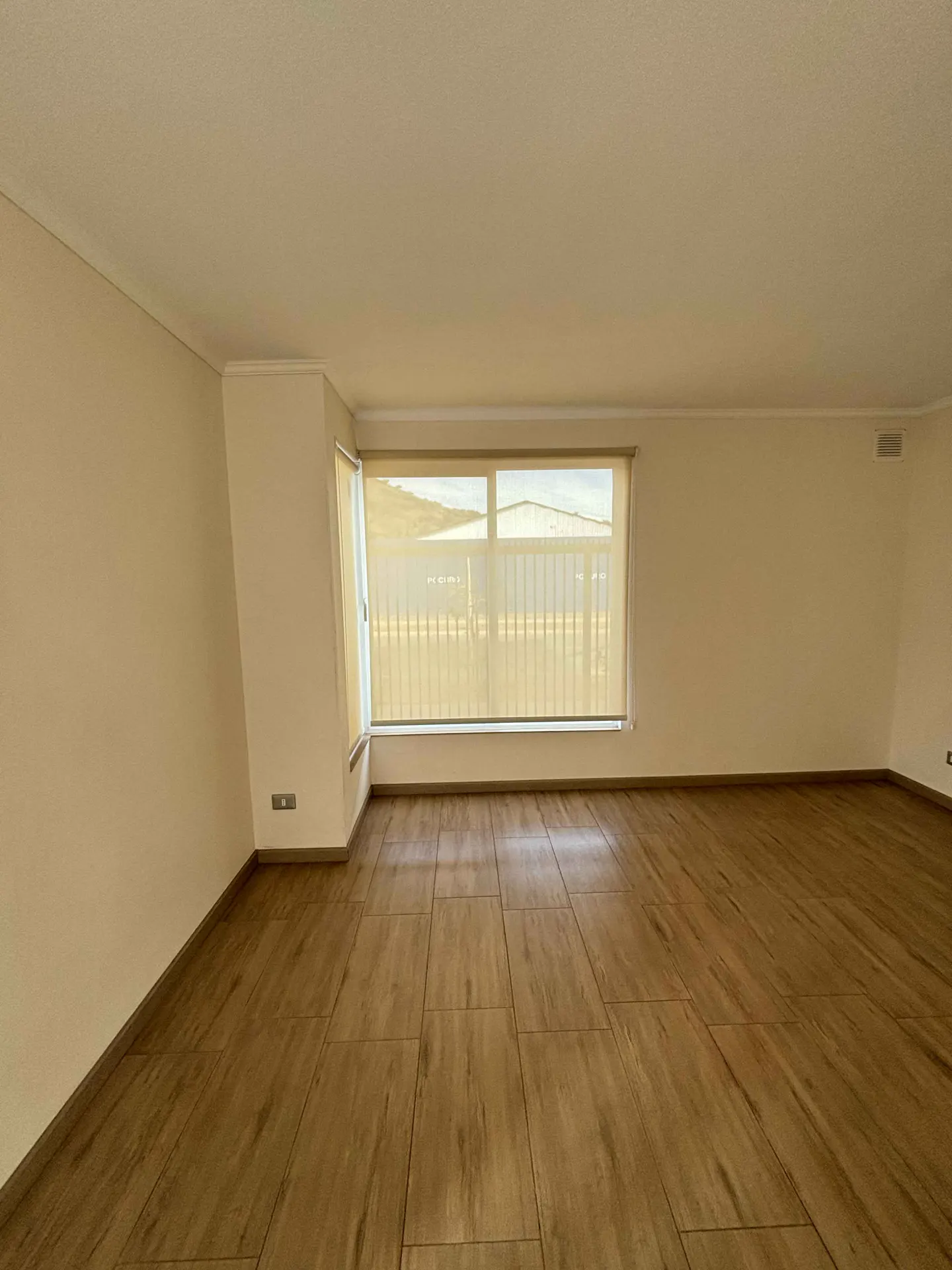 Empty room with wood-look tile floor, beige walls, and a large window with a closed roller shade.