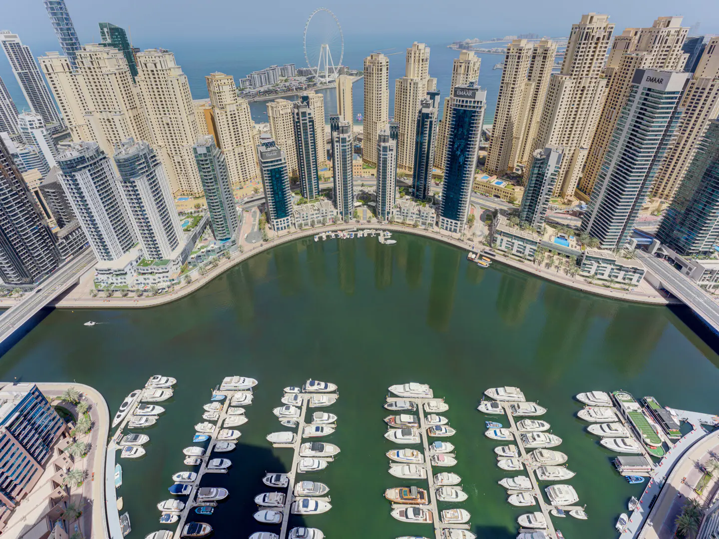 Aerial view of Dubai Marina with skyscrapers, a harbor filled with white yachts, and the Ain Dubai Ferris wheel in the distance.