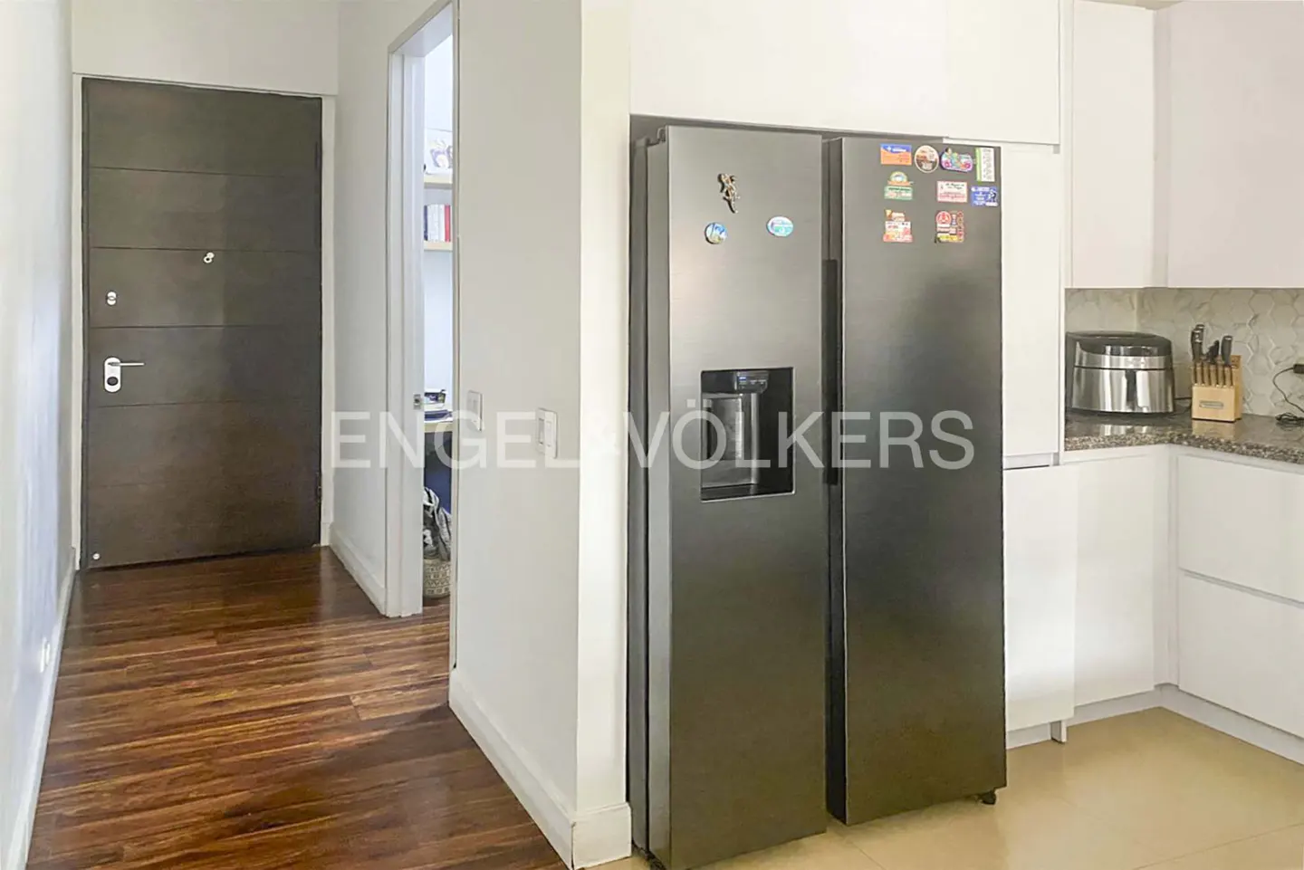 Interior view of a kitchen with a stainless steel refrigerator, white cabinets, and a dark brown door leading to a hallway with wood floors.
