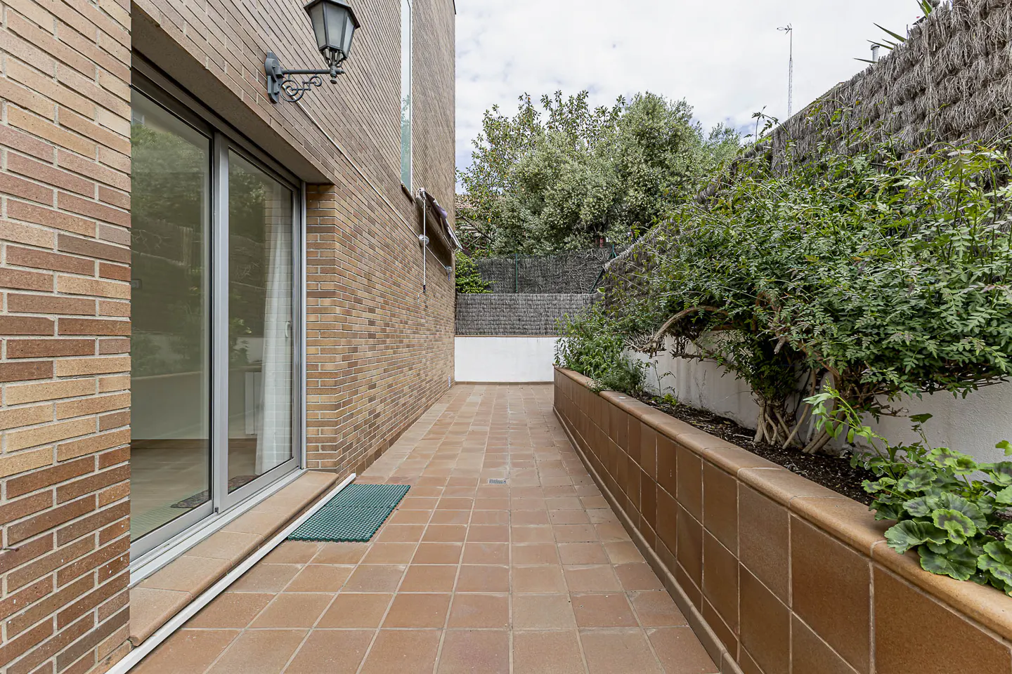 A long, narrow patio with terracotta tiles, a brick wall, and a garden bed with green plants. A sliding glass door leads into the house.