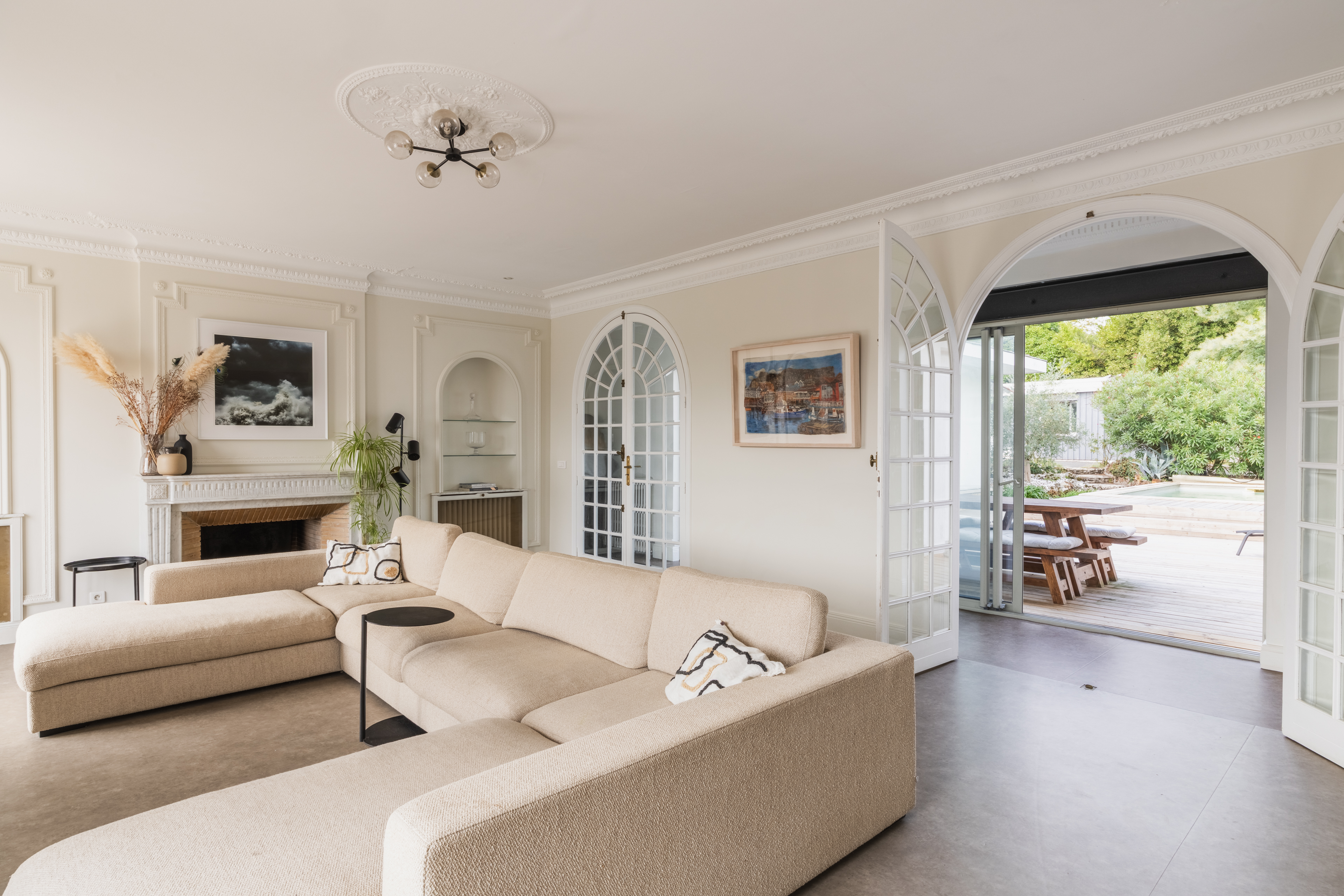 Bright living room with a large beige sectional sofa, fireplace, and arched glass doors opening to a patio with a pool. [AI-generated text]