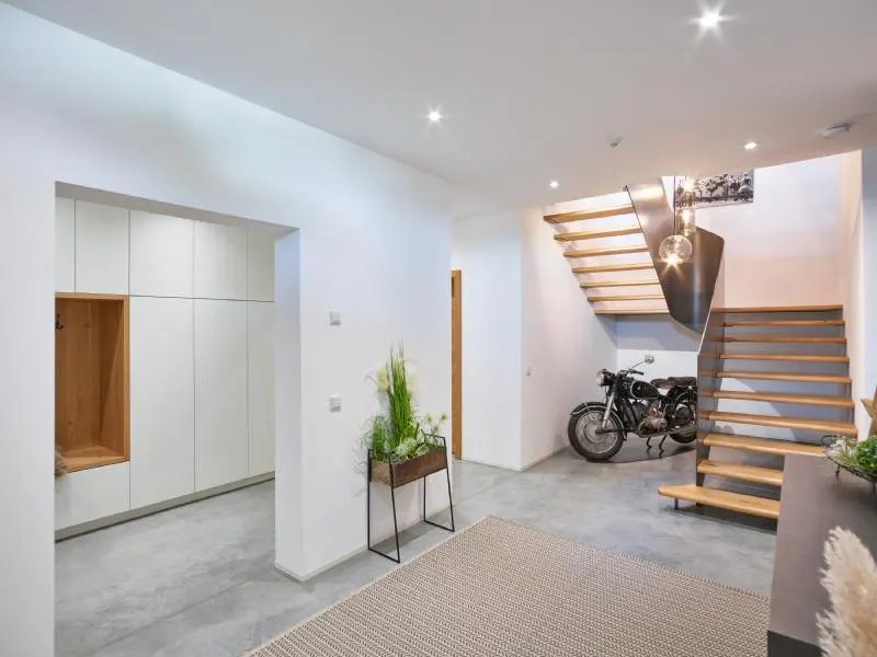 Modern home entryway with white walls, concrete floors, and wooden stairs. A vintage motorcycle sits beneath the stairs. A beige rug is on the floor.