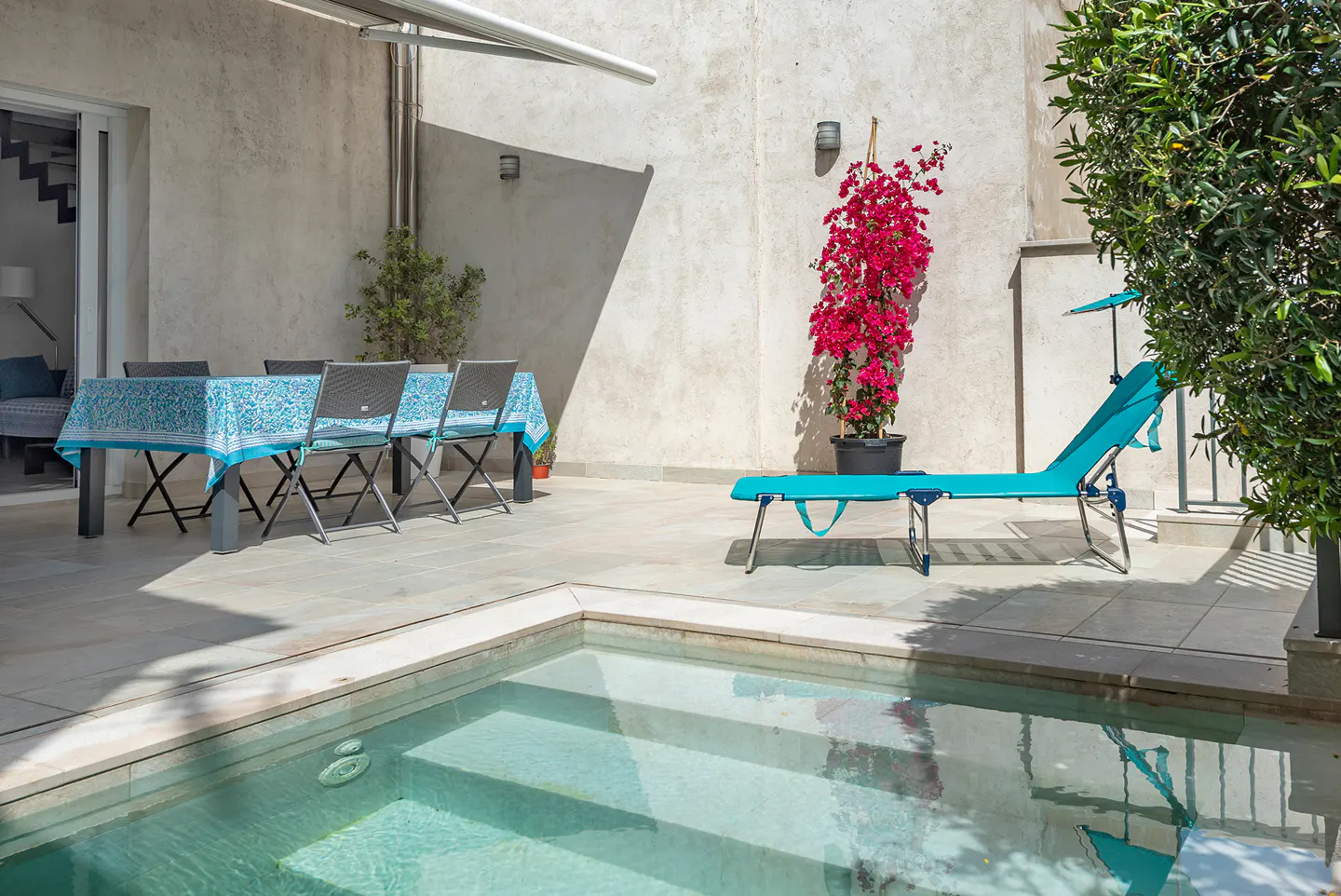 Outdoor patio with a small pool, table with blue tablecloth, chairs, and a blue lounge chair. A bougainvillea plant adds a pop of color.