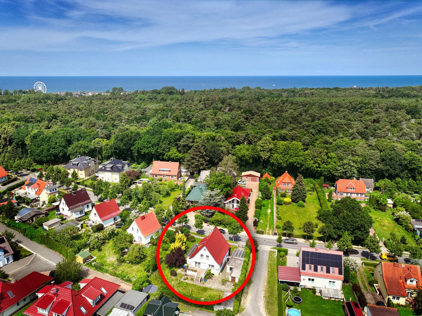 Aerial view of a white house with a red roof, circled in red, in a green neighborhood near a forest and the ocean.