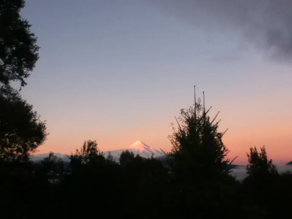 A scenic view of a snow-capped mountain at sunset, framed by silhouetted trees. The sky is a blend of pink and blue hues.