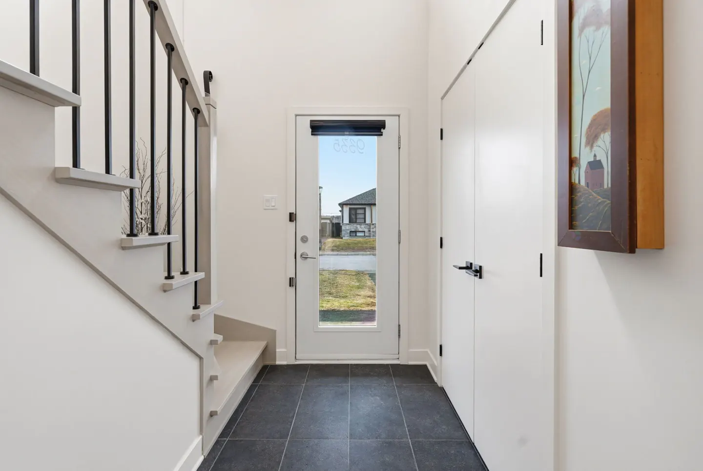 Bright entryway with black tile floor, white walls, and a white door with a view of a house outside. Stairs with black railings are on the left. A painting hangs near a white closet.