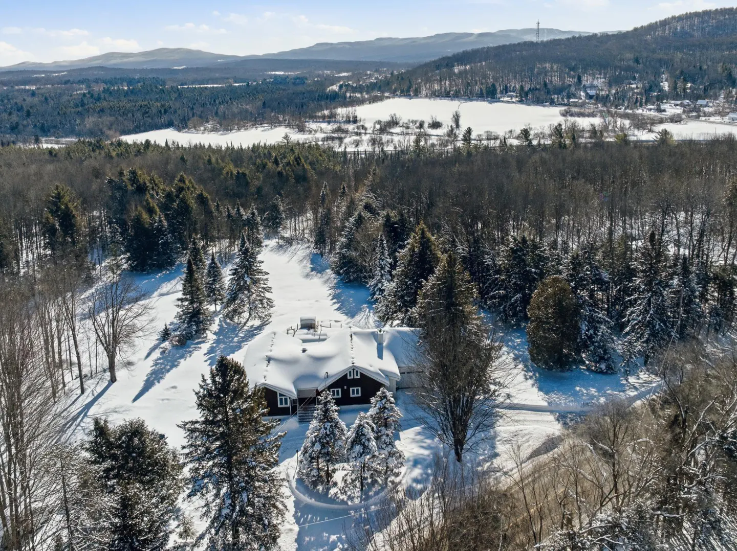 Aerial view of a dark house covered in snow, surrounded by snow-covered trees and a snowy field. Mountains are in the background.