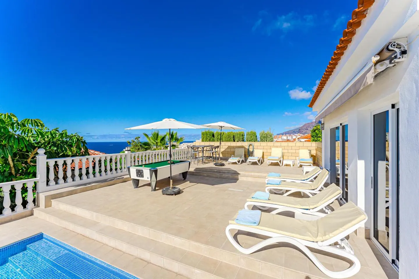Outdoor patio with pool, lounge chairs, and pool table overlooking the ocean under a clear blue sky.