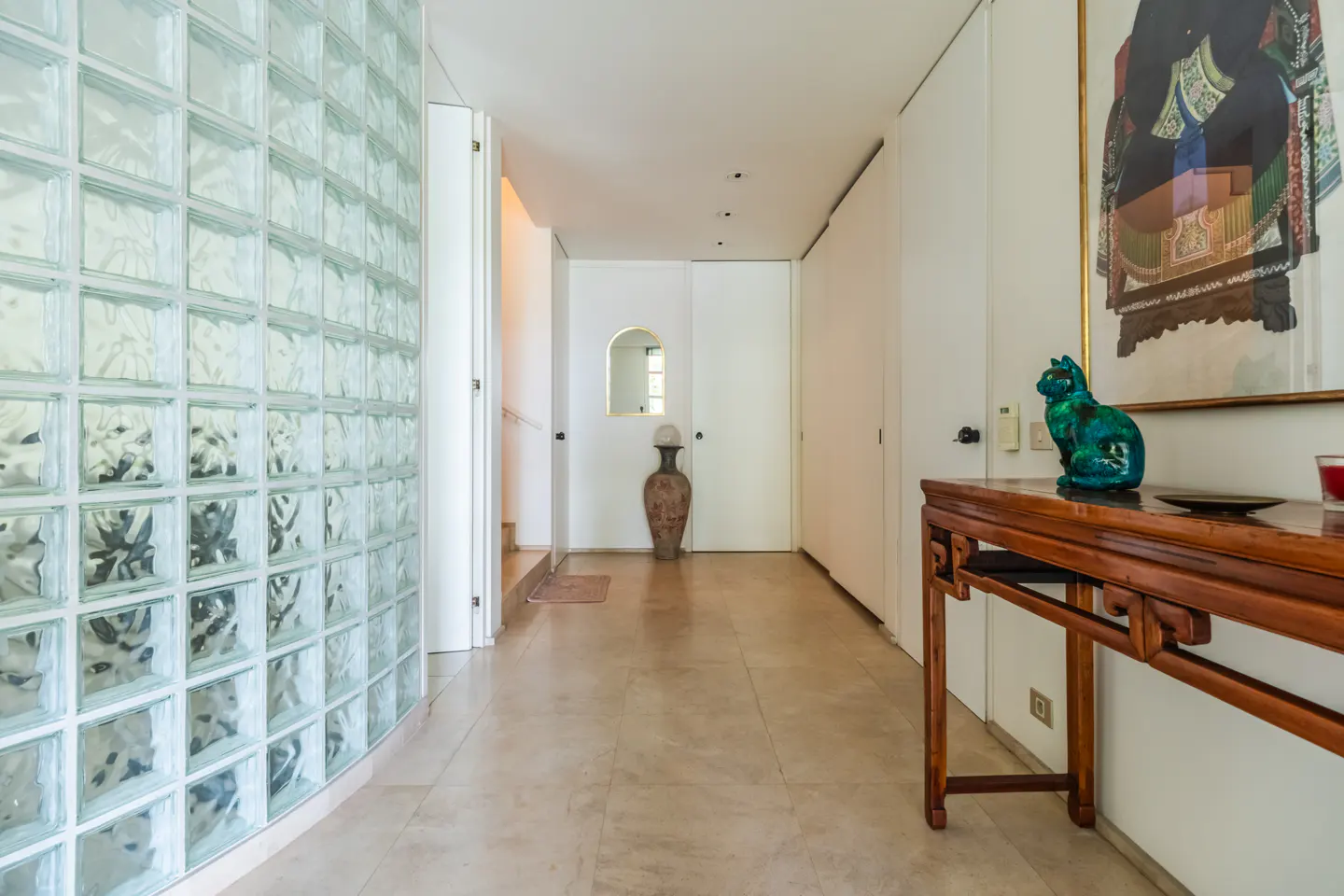Hallway with glass block wall, beige tile floor, and a wooden console table with a green cat statue.