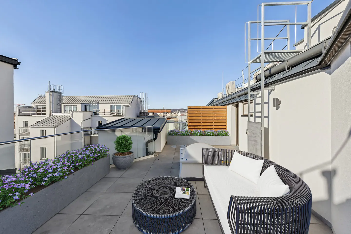 Rooftop patio with a black wicker sofa, round table, and purple flowers in planters. City buildings are visible in the background under a blue sky.
