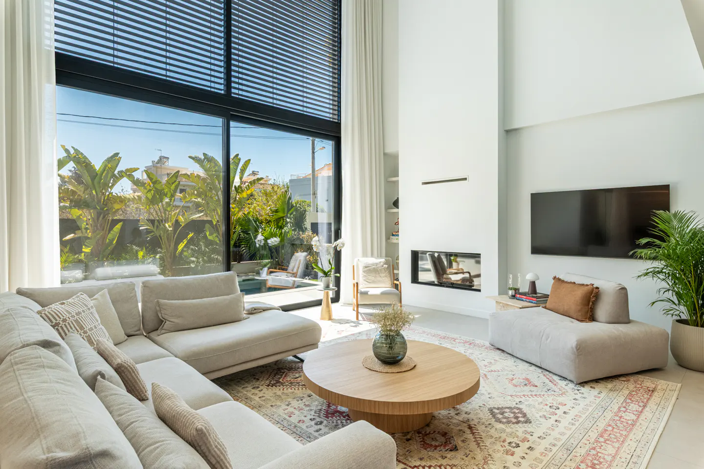Bright living room with a large window, beige sectional sofa, round wood table, and a fireplace under a wall-mounted TV.