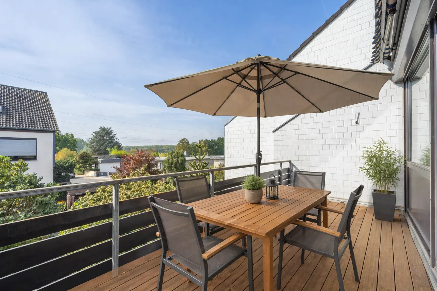 Outdoor patio with a wooden table, chairs, and umbrella on a wooden deck with a black railing. A white brick building is in the background.