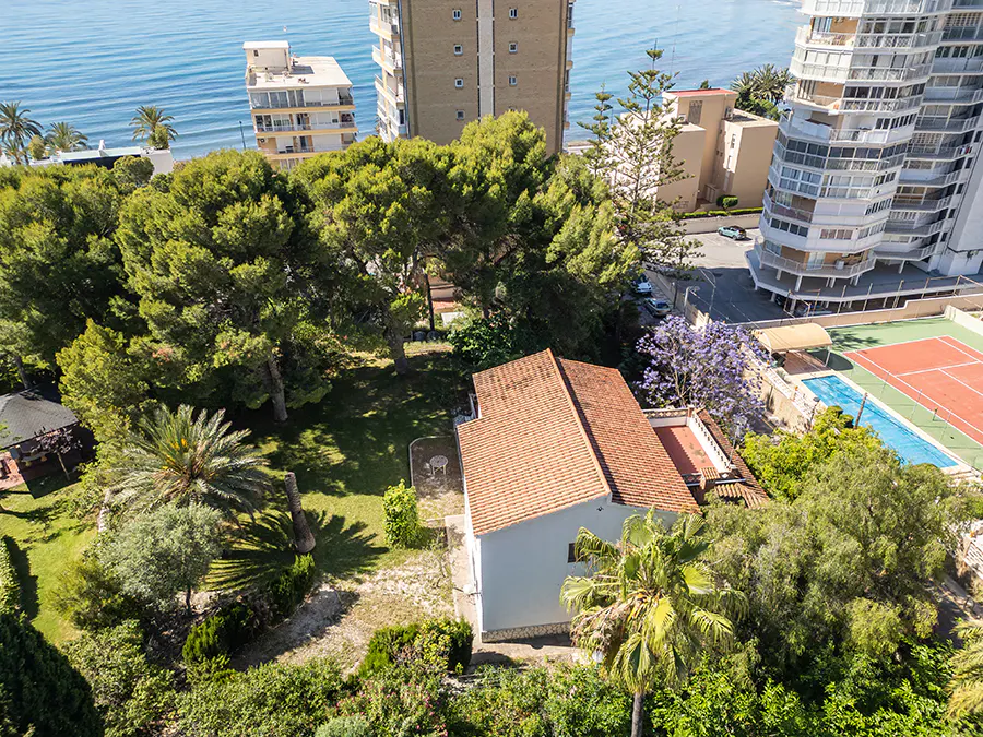 Aerial view of a white house with a red tile roof, surrounded by green trees, near the ocean and tall buildings.