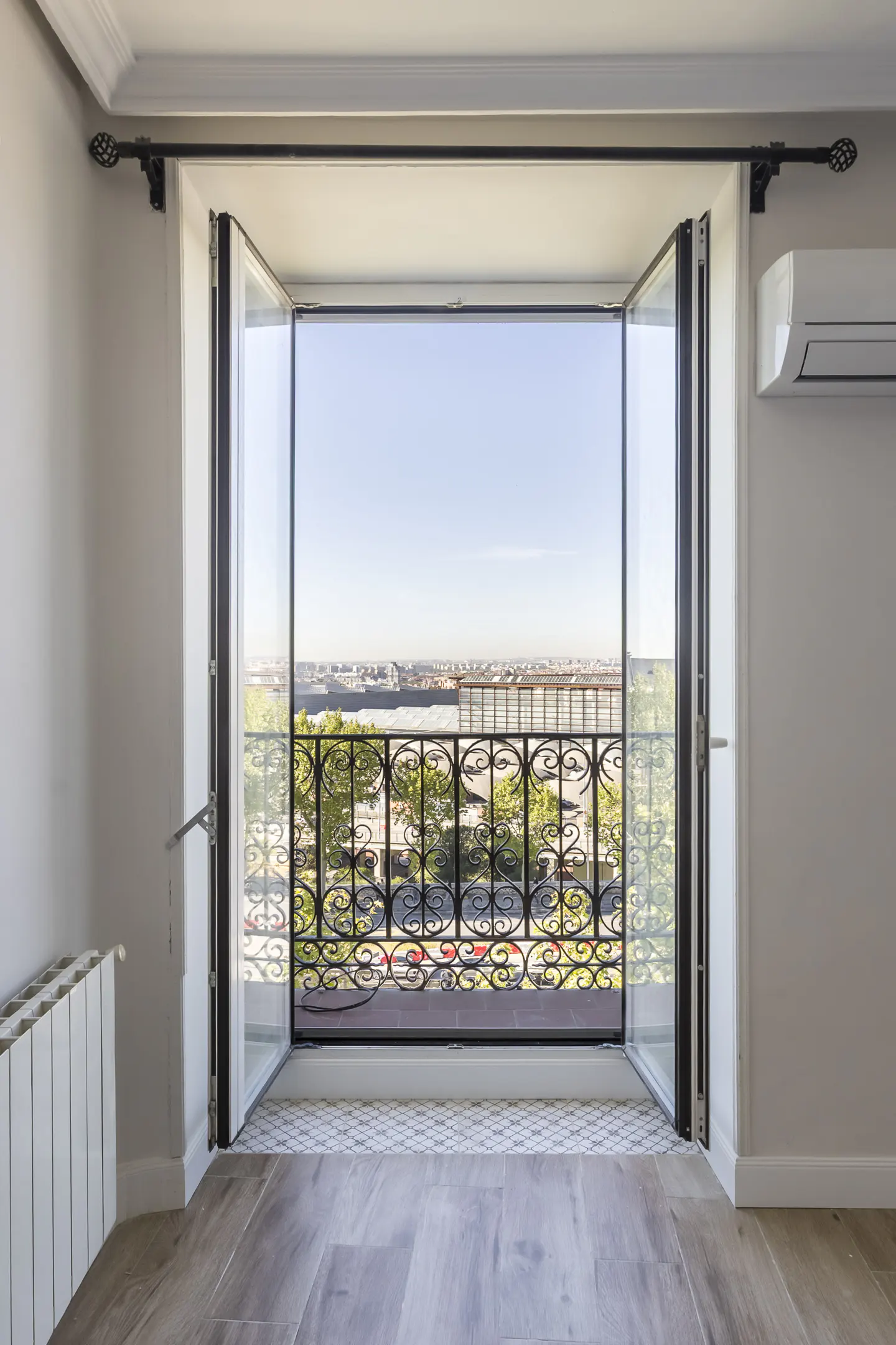 A bright room with open French doors leading to a balcony with a black wrought iron railing and a city view.
