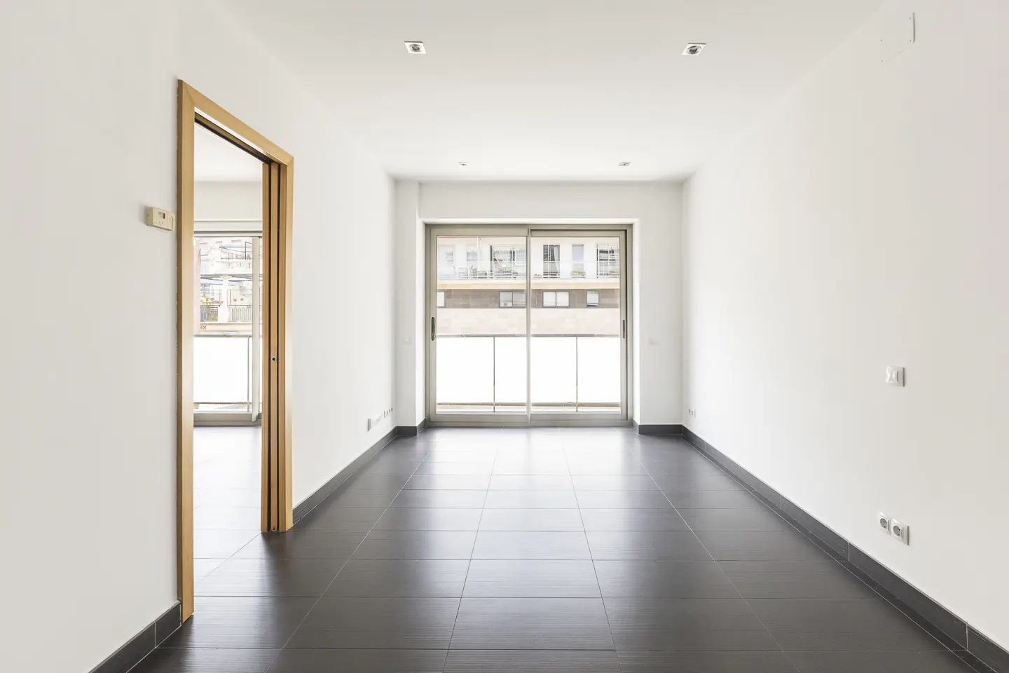 Empty room with white walls, dark tile floor, and a sliding glass door to a balcony. A wooden door frame is on the left.