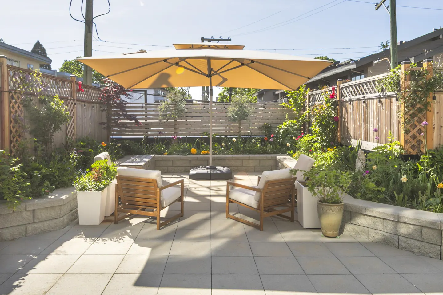 A backyard patio with two wooden chairs, a large tan umbrella, and a stone retaining wall with flower beds.