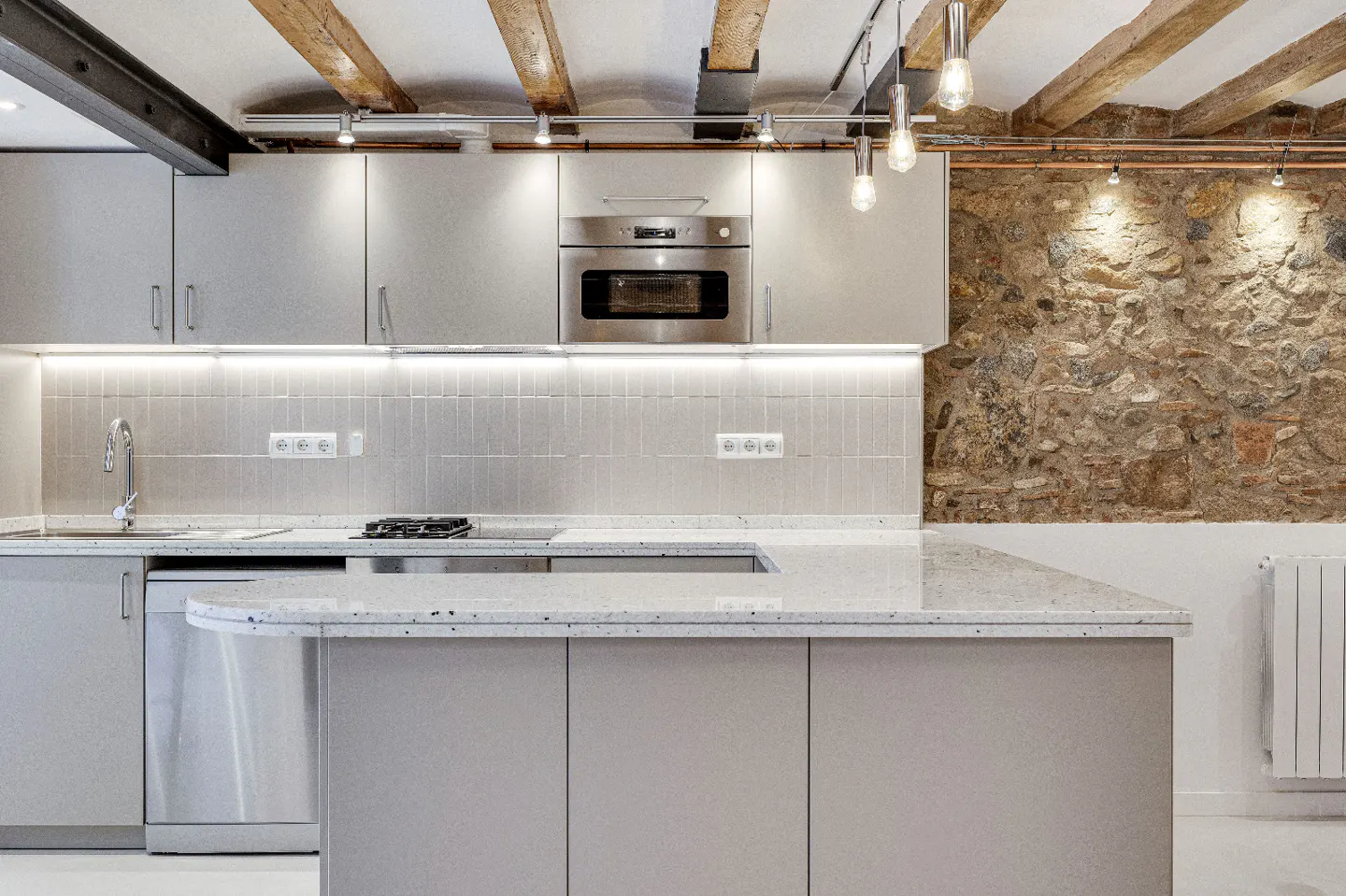 Modern kitchen with gray cabinets, speckled countertops, and stainless steel appliances. Exposed wooden beams and a stone wall add rustic charm.