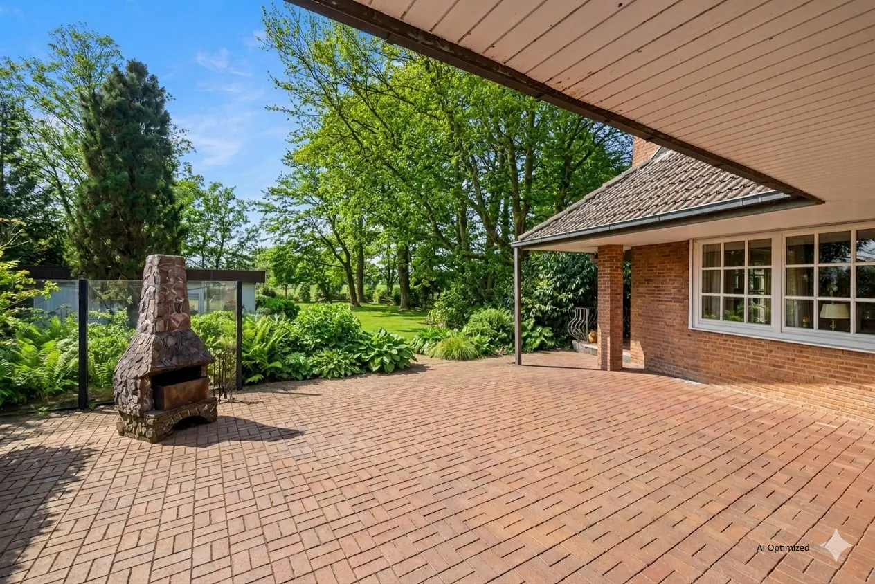 Brick patio with stone fireplace, lush green garden, and brick house under a blue sky.