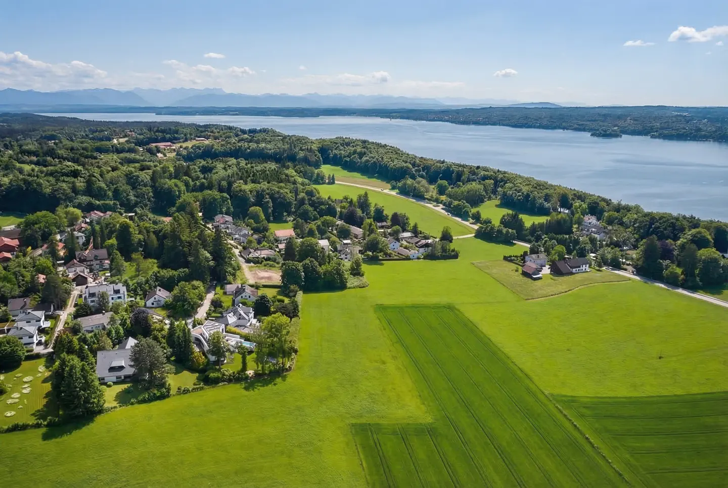 Aerial view of a lakeside community with green fields, trees, and houses under a blue sky. Mountains are visible in the distance.