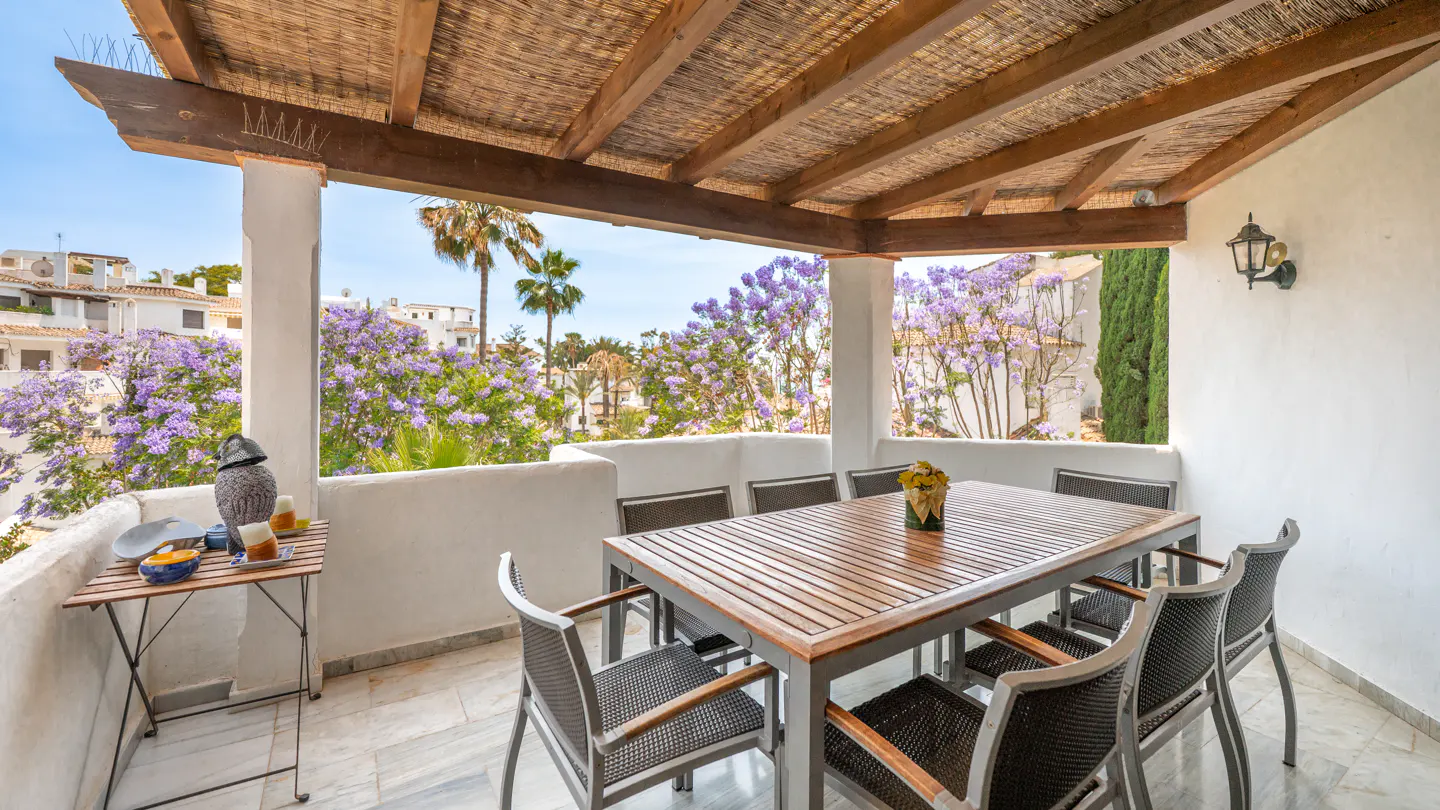 Covered terrace with a wooden dining set, marble floors, and views of blooming purple jacaranda trees and white villas.