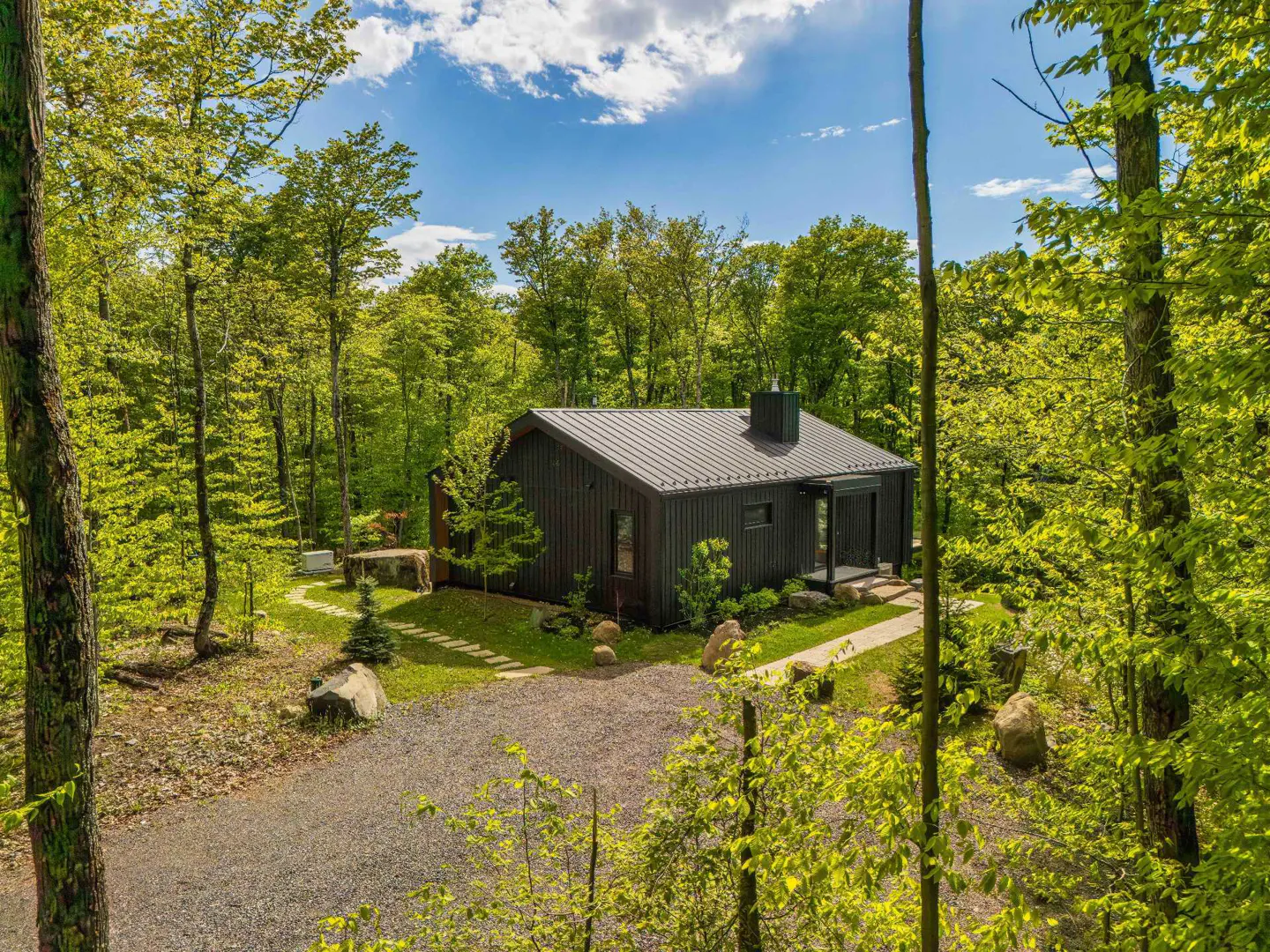 Modern black cabin with a metal roof nestled in a lush, sunlit forest with a gravel driveway and stone path.