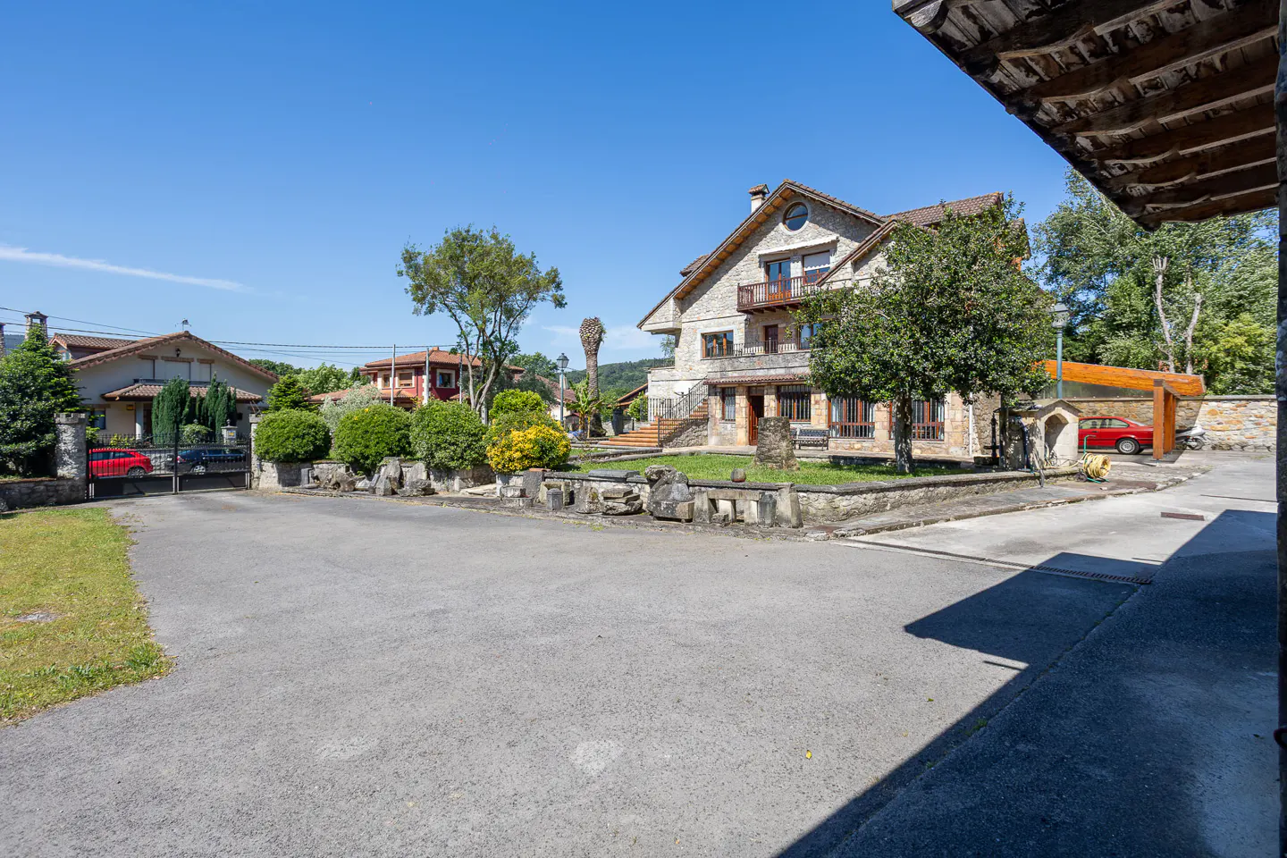 Exterior view of a stone house with a balcony, trees, and a driveway on a sunny day.