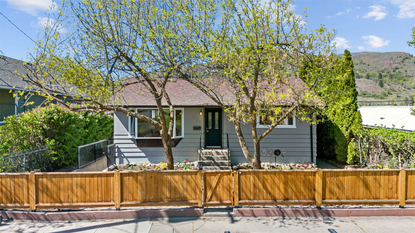 A gray house with a brown roof, a dark green door, and a wooden fence in front. Trees are in the yard.
