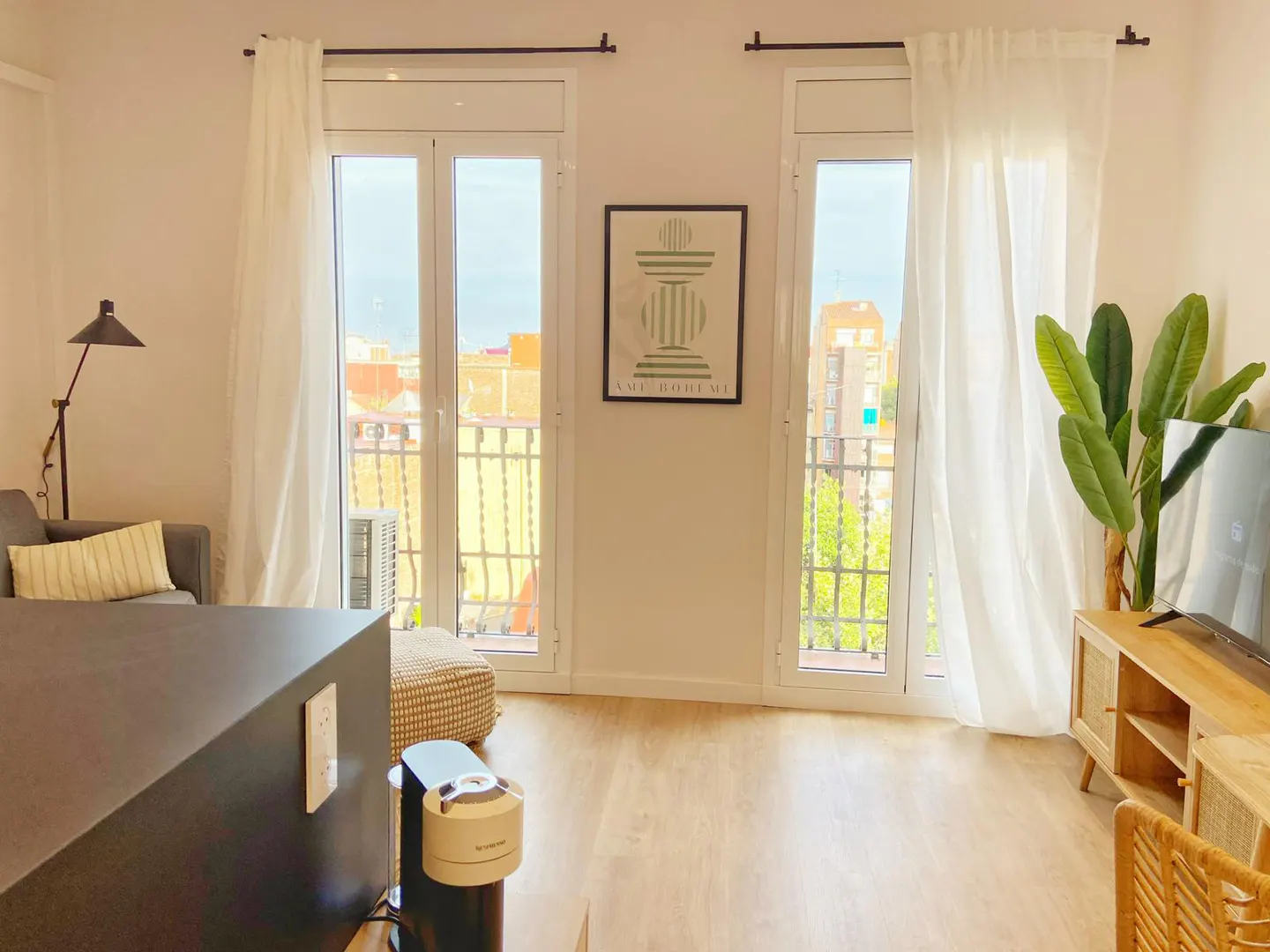 Bright living room with hardwood floors, white curtains, and balcony doors. A gray sofa, TV, and plant add modern touches.