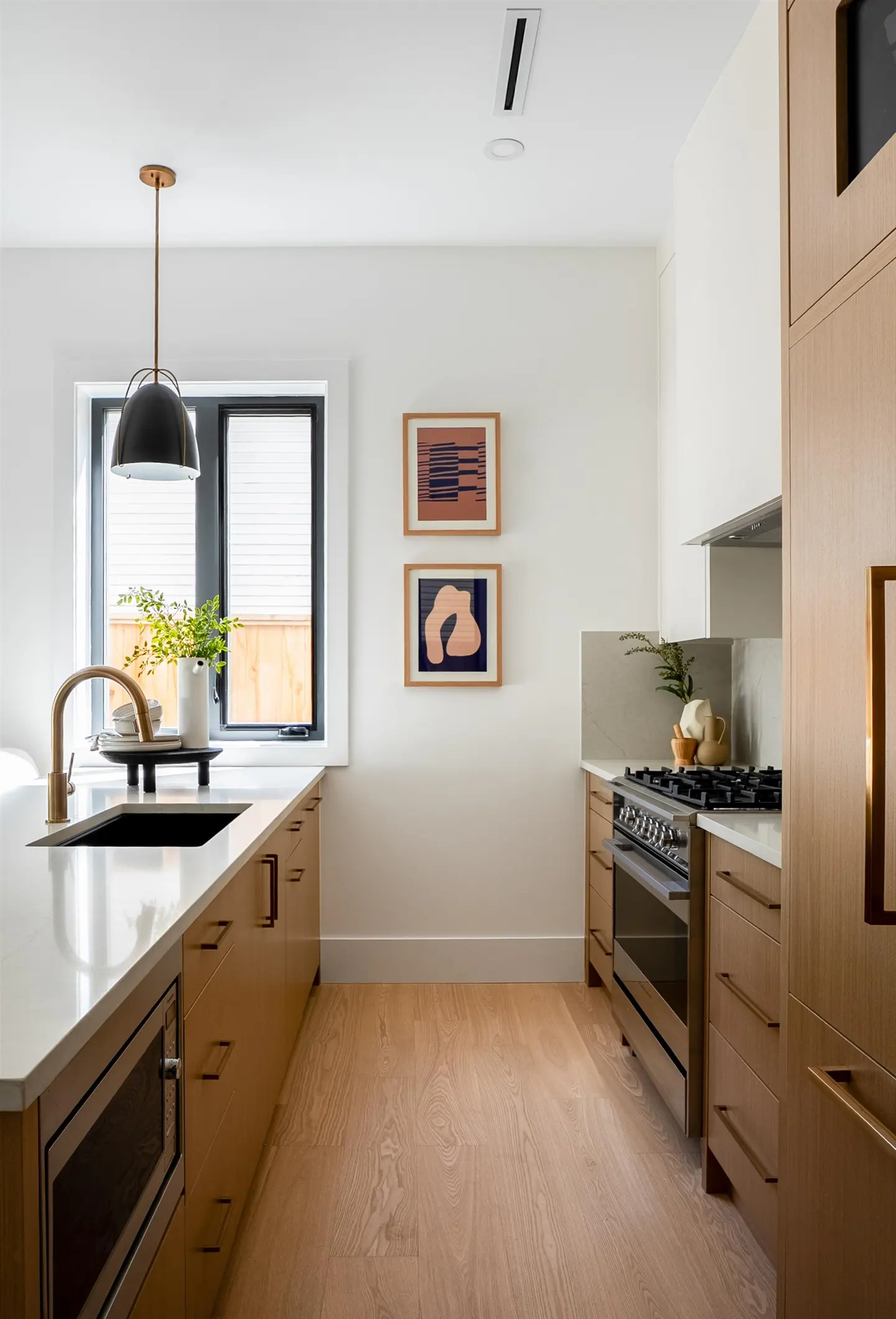 A modern kitchen with light wood cabinets, white countertops, and stainless steel appliances. Two framed art pieces hang on the white wall.