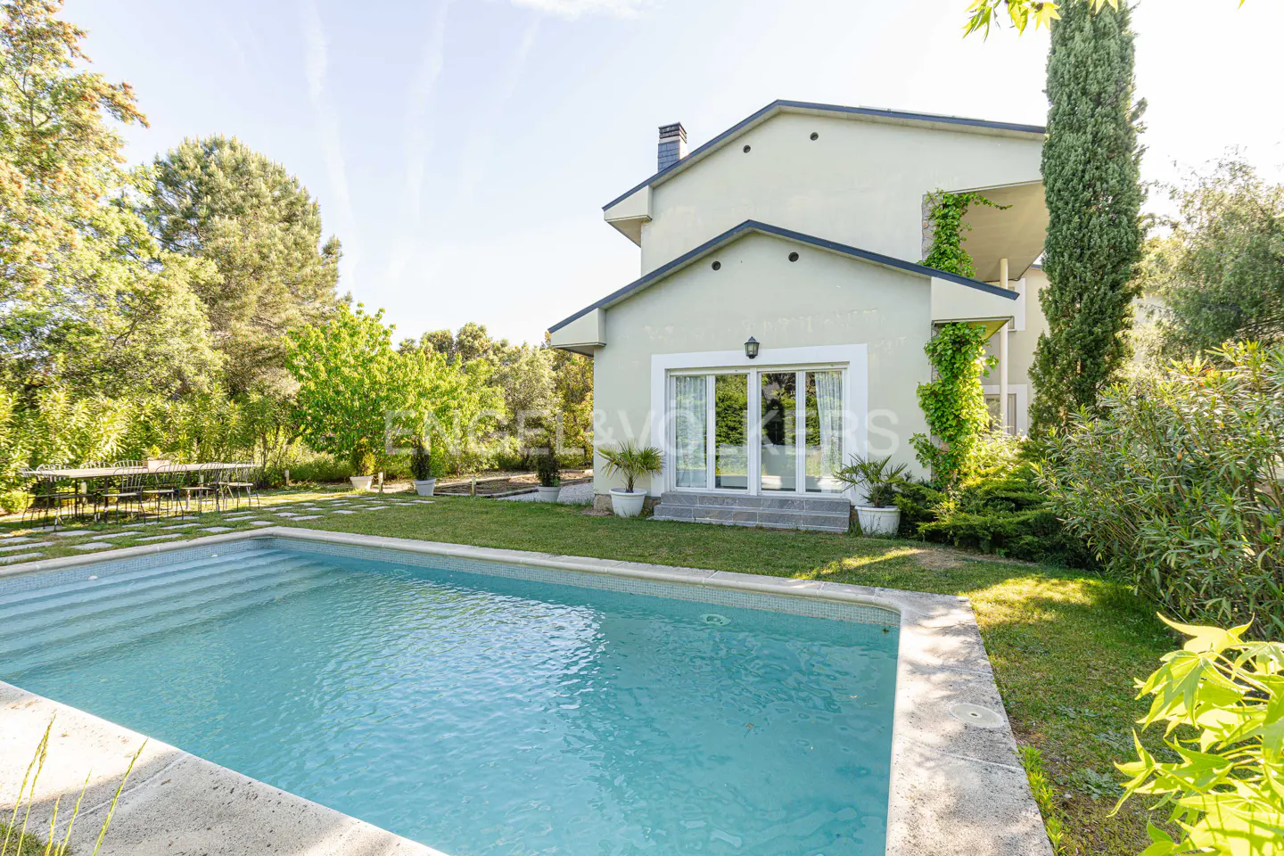 Exterior view of a two-story house with a pool, green lawn, and trees under a sunny sky.