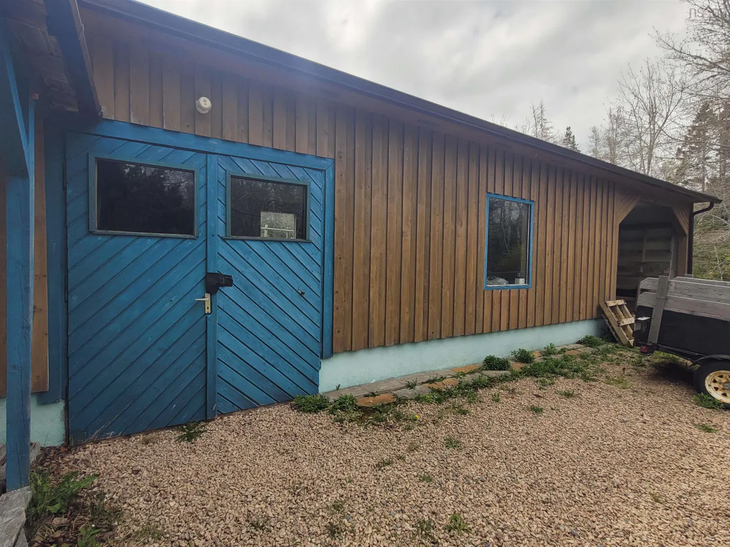 Exterior of a brown wooden building with blue doors and trim, gravel ground, and a trailer parked nearby.
