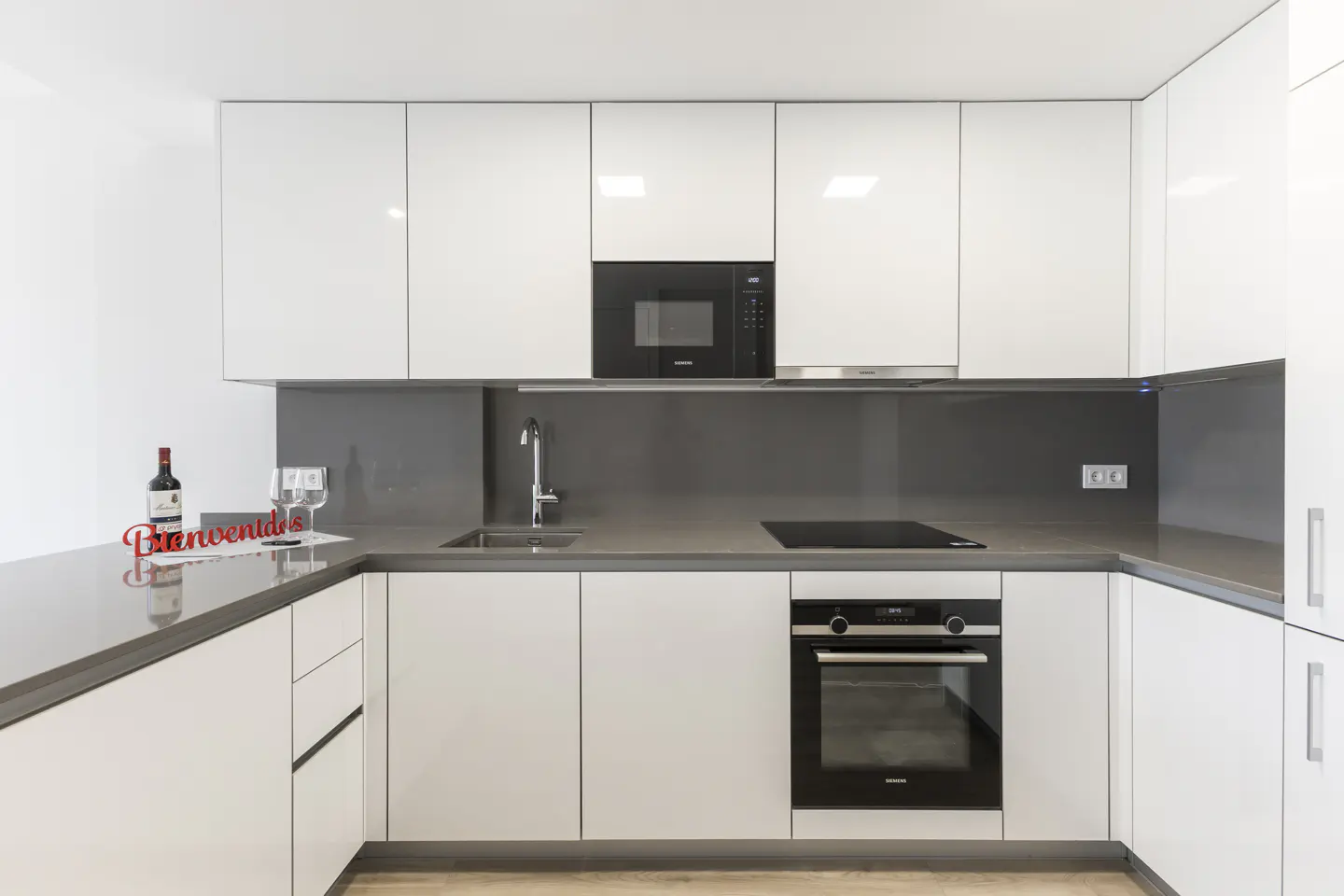Modern kitchen with white glossy cabinets, grey countertops, built-in black appliances, and a wine bottle on the counter.