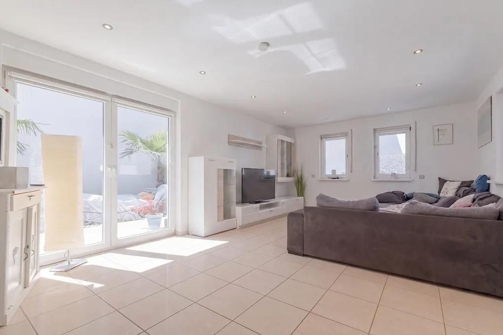 Bright living room with tile floor, white walls, and a large gray sectional sofa. Sliding glass doors lead to a patio with a palm tree.